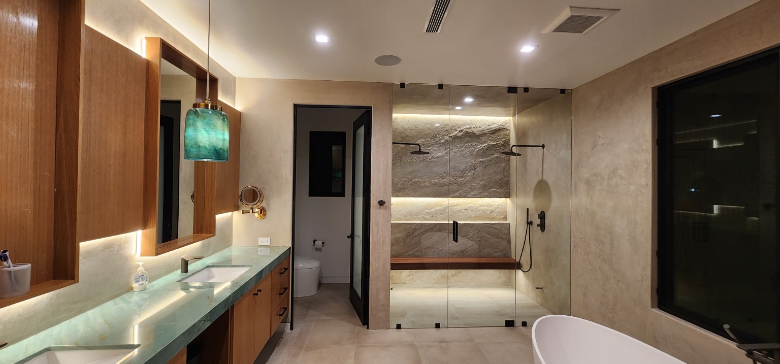 Wood vanity with green countertop, framed mirrors, pendant lighting, and illuminated shower beyond